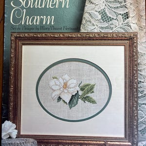 Può includere: Un ricamo a punto croce incorniciato di un fiore bianco con foglie verdi. La cornice è marrone e dorata. L'immagine è su un libro intitolato "Southern Charm" di Leisure Arts.