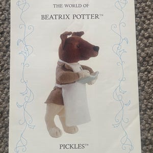 Puede incluir: Una página impresa con un personaje de perro tejido de "The World of Beatrix Potter". El perro lleva una chaqueta marrón, un delantal blanco y sostiene un plato pequeño. El título y el nombre del personaje se muestran.