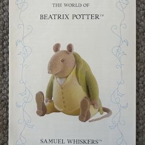Puede incluir: Una tarjeta impresa con el texto THE WORLD OF BEATRIX POTTER y SAMUEL WHISKERS. En el centro aparece una rata tejida con un chaleco amarillo y una chaqueta verde, enmarcada por un fino borde decorativo azul sobre un fondo blanco.
