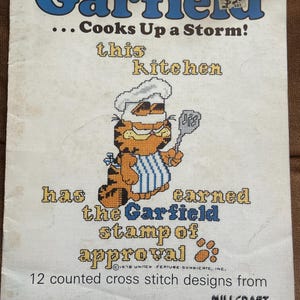 May include: A Garfield cross stitch pattern booklet titled Garfield... Cooks Up a Storm! featuring a cross-stitched illustration of Garfield wearing a chef hat and blue striped apron while holding a spatula, with the text this kitchen has earned the Garfield stamp of approval and 12 counted cross stitch designs from Millcraft, Inc.