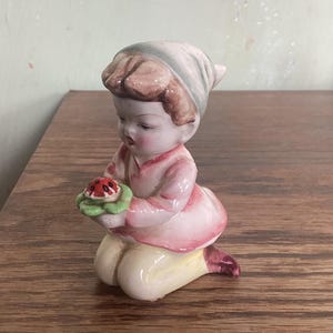 May include: A ceramic figurine of a girl kneeling and holding a ladybug in her hand. She is wearing a pink dress and a white hat. The figurine is painted in shades of pink, white, green, and red.