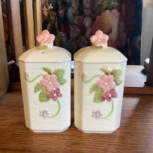 Puede incluir: Un par de saleros y pimenteros de cerámica blanquecina. Cada salero y pimentero presenta un diseño floral con flores rosas y moradas, hojas verdes y una flor rosa en la parte superior. Los saleros tienen un patrón geométrico texturizado.