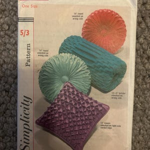 Könnte beinhalten: Vintage Simplicity-Schnittmusterumschlag mit Illustrationen von Smokkissen in verschiedenen Formen und Farben. Der Umschlag zeigt ein rotes, blaugrünes und lila Kissen-Design. Der Text auf dem Umschlag lautet "5244 One Size" und "Simplicity Pattern".