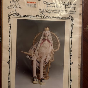May include: A doll kit for a "Domestic Goddess Fertility Fairy" doll. The doll is seated in a wicker chair and wears a crown and sandals. The kit includes instructions for making 23-inch and 10-inch cloth dolls. The package is labeled "Magic Threads."