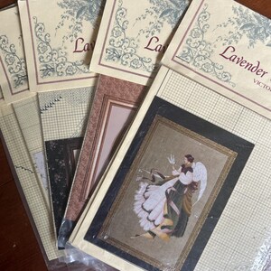 May include: A collection of stacked paper craft patterns featuring cream-colored backgrounds with grid lines and decorative floral borders. The top pattern displays an illustration of an angel with wings, while the text Lavender reads clearly.