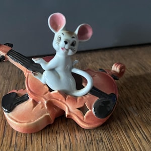 May include: A ceramic figurine of a white mouse with pink ears playing a violin. The violin is a reddish-orange color with black details. The mouse is smiling, and the figurine is set against a neutral background.