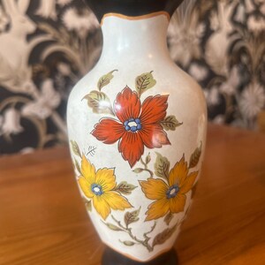 Może przedstawiać: Ozdobny ceramiczny wazon z białą podstawą, czarną obręczą i podstawą oraz kolorowym kwiatowym wzorem. Kwiaty są pomarańczowe i żółte z niebieskimi środkami i zielonymi liśćmi. Wazon ma około 20 cm wysokości.