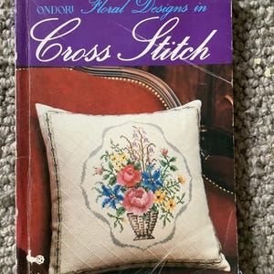 Op de afbeelding: Een boek getiteld "Floral Designs in Cross Stitch" met een paarse omslag. De omslag toont een kruissteek kussen met een bloemenmand ontwerp in roze, blauw en geel. Het boek ligt op een rode ondergrond.