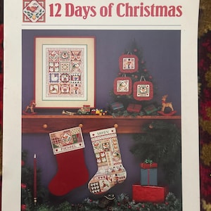 May include: A Dimensions cross-stitch pattern booklet titled A Classic Christmas Collection 12 Days of Christmas. The cover features a framed sampler, three ornaments, and two stockings, one red and one patterned, hanging on a wooden mantelpiece.
