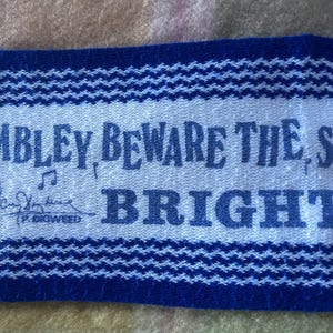 May include: A rectangular fabric patch with a light gray center and dark blue borders featuring a woven texture. The text reads "MBLEY, BEWARE THE S" and "BRIGHT" with a musical note symbol and a signature above the text "P. DIGWEED".