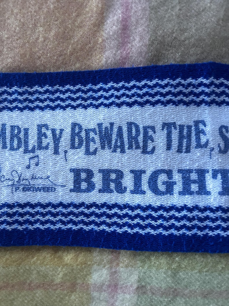 May include: A rectangular fabric patch with a light gray center and dark blue borders featuring a woven texture. The text reads "MBLEY, BEWARE THE S" and "BRIGHT" with a musical note symbol and a signature above the text "P. DIGWEED".