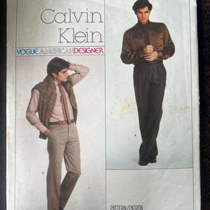 Könnte beinhalten: Eine Vogue American Designer Schnittmusterhülle für das Calvin Klein Design 2256. Die Vorderseite zeigt Illustrationen von zwei Outfits: eines mit einem braunen Hemd und dunkler Hose und eines mit einem hellen Hemd, brauner Strickweste und Hose.