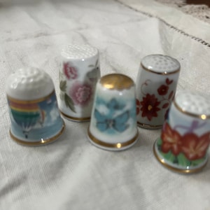 Puede incluir: Cinco dedal de porcelana con diferentes diseños. Los dedales son blancos con ribetes dorados y presentan motivos florales y de mariposas coloridos.