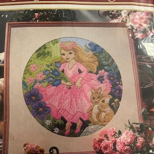 Può includere: Un kit per ricamo Semco Long Stitch Originals con un disegno circolare a punto croce di una persona con capelli biondi in un abito rosa che tiene fiori blu, accompagnata da due conigli marroni in un giardino con fiori rosa e viola.