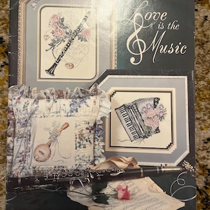 Può includere: Una copertina di libro con il testo "Love is the Music" e illustrazioni di strumenti musicali. La copertina presenta un clarinetto, un banjo e una fisarmonica, ciascuno accompagnato da composizioni floreali. Il libro è etichettato "book 78" e proviene dalla "Stoney Creek Collection".