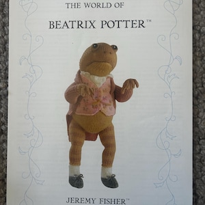 Puede incluir: Una tarjeta impresa con una figura de rana tejida de Jeremy Fisher de The World of Beatrix Potter. La rana lleva un chaleco floral rosa, una chaqueta marrón, calcetines blancos y zapatos oscuros. El texto dice THE WORLD OF BEATRIX POTTER y JEREMY FISHER.
