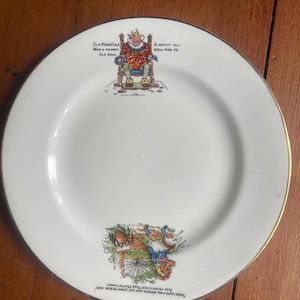 May include: White ceramic plate with a gold rim featuring illustrations and text. The top illustration depicts a king, and the bottom shows a spider and a girl. Text includes "Old King Cole" and "A Merry Old Soul".