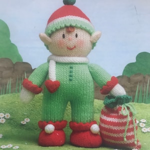 Pode incluir: Uma boneca elfo de tricô vestindo um macacão verde, sapatos vermelhos com pompons brancos e um gorro vermelho e verde com um pompom. A boneca tem um cachecol branco com um coração vermelho, pele creme e segura uma bolsa listrada vermelha e branca.