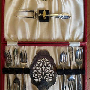 May include: A vintage silverware set in a red case. The set includes a carving fork, serving utensils, and several forks. The case is lined with cream-colored fabric. The silverware has decorative handles and is arranged neatly.