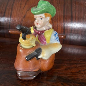 Puede incluir: Una figurita de cerámica de un vaquero con un sombrero verde, un pañuelo rojo y una camisa azul y amarilla. Sostiene una pistola en su mano derecha y una botella en su mano izquierda.