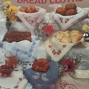Può includere: Immagine di canovacci per il pane ricamati a punto croce con motivi floreali e di cigni. I canovacci sono esposti con prodotti da forno, fiori e un bicchiere di liquido. Il testo "Sam Hawkins Designs Cross Stitch for BREAD CLOTHS" è visibile.