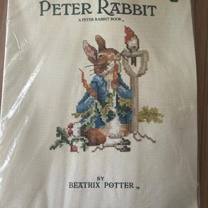 Peut inclure: Un livret de modèles de point de croix intitulé The Tale of Peter Rabbit par Beatrix Potter, Book 549. La couverture présente une illustration au point de croix de Pierre Lapin en veste bleue tenant une carotte près d'une pelle sur une grille crème.