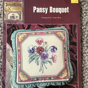 Può includere: Un libretto di schemi a punto croce intitolato "Pansy Bouquet" di Linda Bird. Il disegno presenta una composizione floreale di viole nei toni del viola, rosso e rosa. La dimensione del cuscino finito è di 40,6 cm x 40,6 cm.