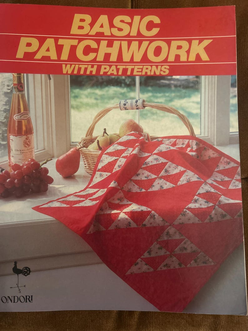 Może przedstawiać: Książka w miękkiej oprawie zatytułowana BASIC PATCHWORK WITH PATTERNS z czerwono-białym geometrycznym quiltem na okładce. Obraz na okładce przedstawia czerwono-biały patchworkowy quilt, wiklinowy kosz z gruszkami, butelkę wina i winogrona.