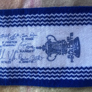 May include: A blue and white woven scarf featuring the FA Cup trophy graphic, the text 1983 FA CUP FINALISTS, and several printed signatures including G. Ryan, S. Foster, G. Ramsey, and G. Smith, with a textured knit pattern throughout.