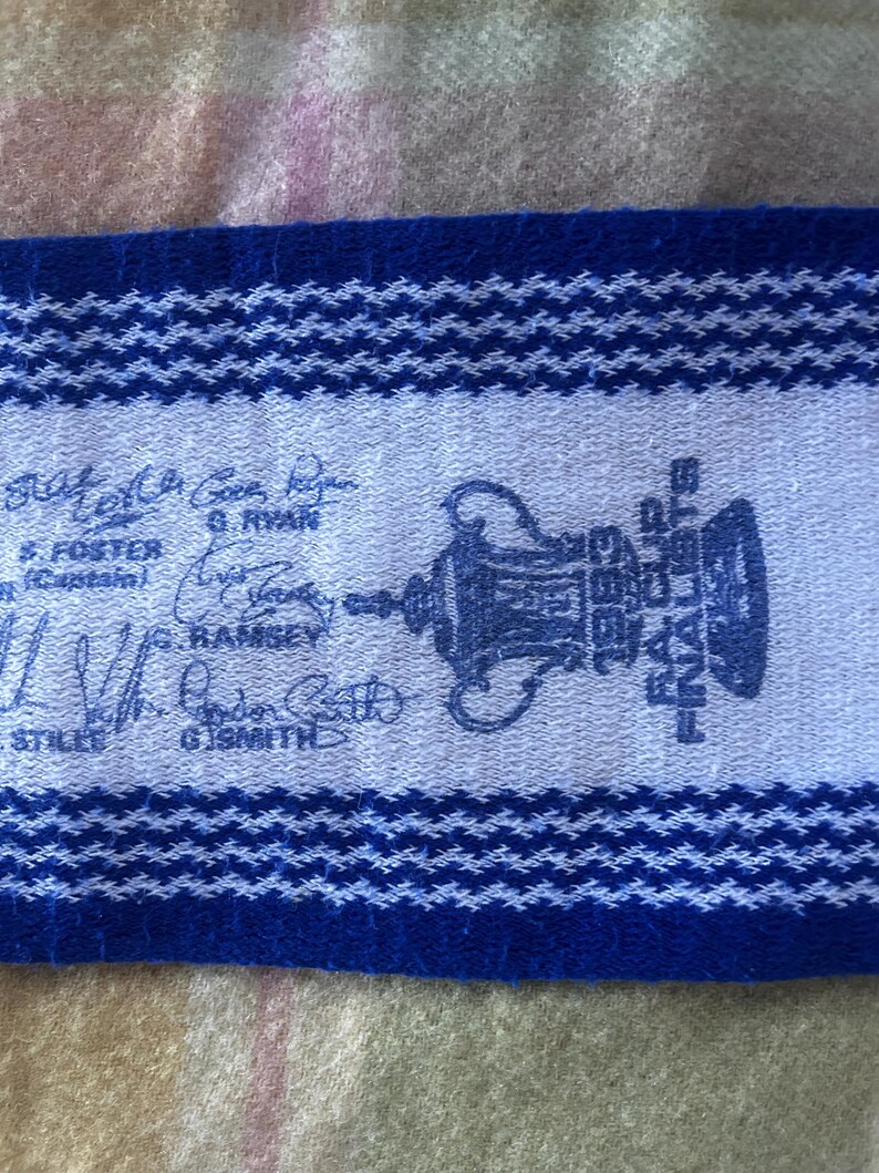 May include: A blue and white woven scarf featuring the FA Cup trophy graphic, the text 1983 FA CUP FINALISTS, and several printed signatures including G. Ryan, S. Foster, G. Ramsey, and G. Smith, with a textured knit pattern throughout.