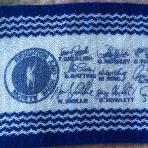 May include: A blue and white woven fabric strip featuring a circular logo with a silhouette of a person, surrounded by text. The strip includes multiple handwritten signatures and printed names such as T. Grealish, G. Mosley, S. Batting, N. Ring, N. Smillie, and G. Howlett.
