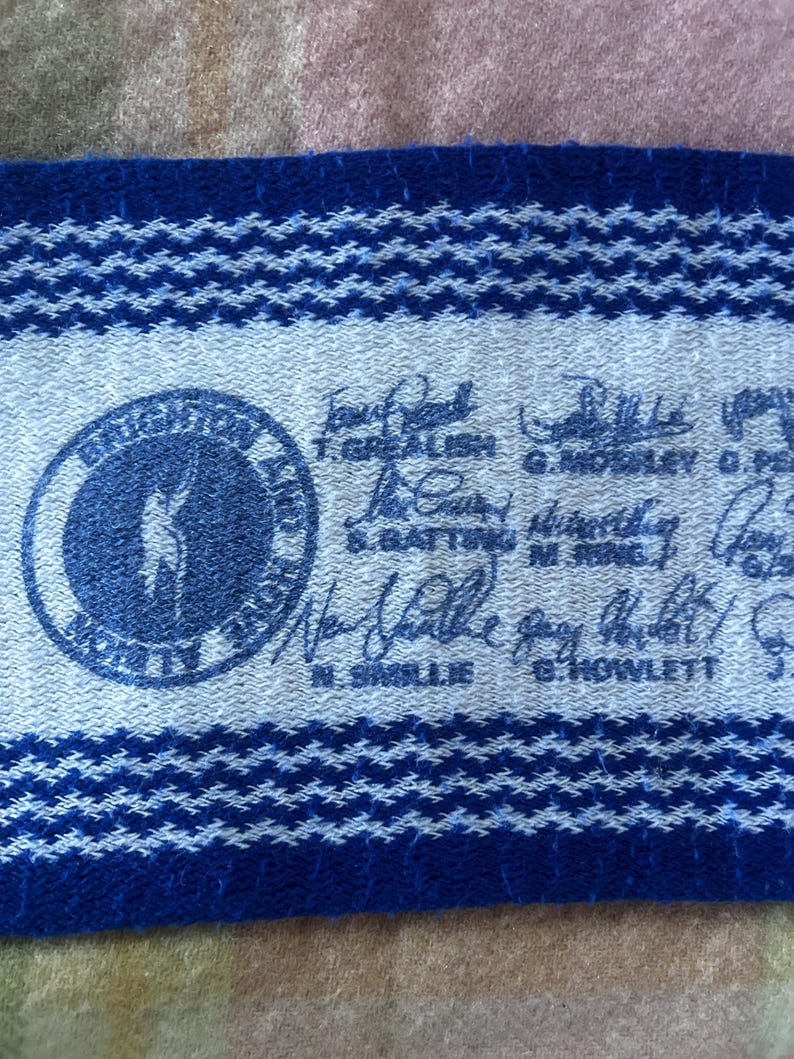 May include: A blue and white woven fabric strip featuring a circular logo with a silhouette of a person, surrounded by text. The strip includes multiple handwritten signatures and printed names such as T. Grealish, G. Mosley, S. Batting, N. Ring, N. Smillie, and G. Howlett.