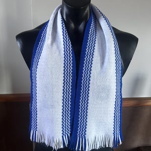 May include: A knitted scarf featuring a white base with two vertical blue stripes. The stripes have a textured, zigzag pattern running through the center. The scarf ends are finished with white and blue fringe detailing along the bottom edges.