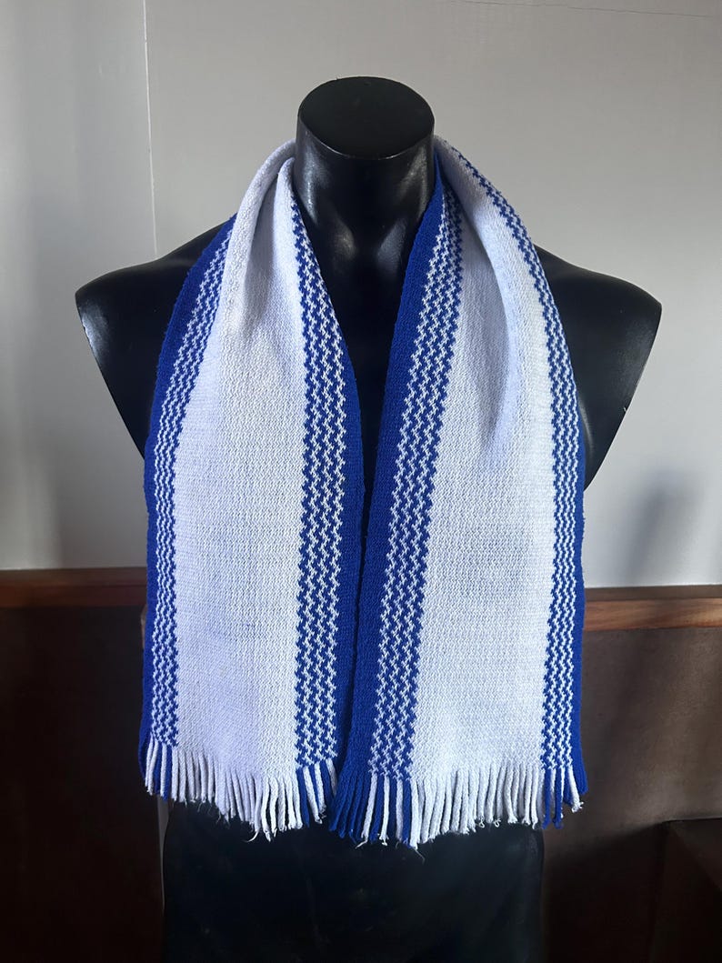 May include: A knitted scarf featuring a white base with two vertical blue stripes. The stripes have a textured, zigzag pattern running through the center. The scarf ends are finished with white and blue fringe detailing along the bottom edges.