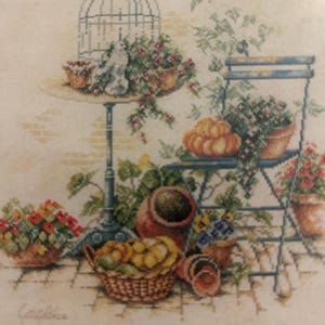May include: A cross-stitch depiction of a garden scene with a birdcage, table, and folding chair. The scene includes potted flowers, a basket of fruit, and a pumpkin. The color palette is soft, with muted tones of green, pink, and yellow.