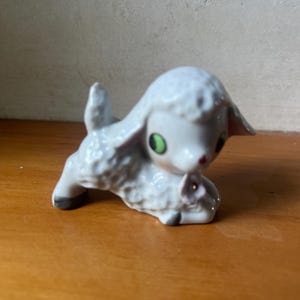 Puede incluir: Pequeña figura de cordero de cerámica blanca, con ojos verdes y nariz rosa. El cordero está tumbado con las patas delanteras cruzadas y las orejas levantadas. La figura tiene un acabado brillante y es un objeto decorativo.