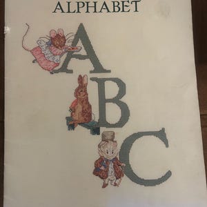 May include: A white cross-stitch pattern booklet titled Beatrix Potter Alphabet with the letters A, B, and C in dark gray. The letter A features a mouse in a pink dress, B shows a rabbit on a bench, and C depicts a character in a hat and coat.