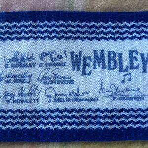 May include: A blue and white woven fabric strip featuring the text WEMBLEY and a musical note icon. The fabric displays multiple printed signatures and names including G. MOSLEY, G. PEARCE, M. RINK, G. STEVENS, G. HOWLETT, J. MELIA, and P. GIGWEED.