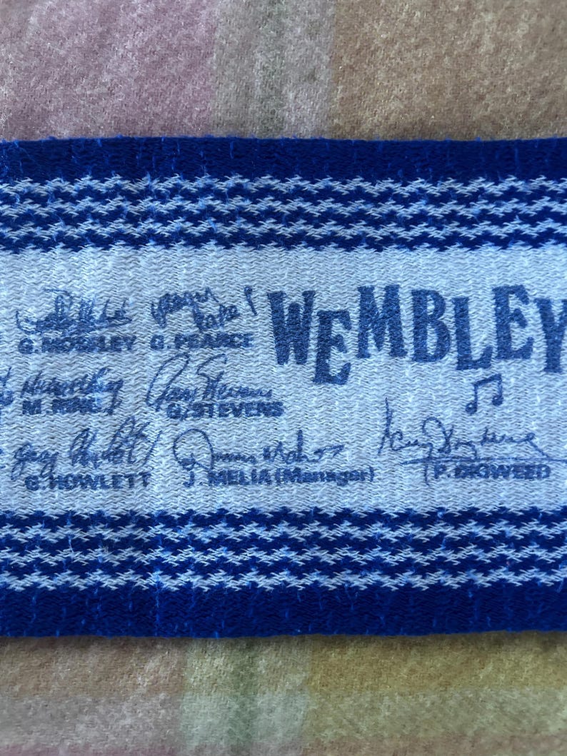 May include: A blue and white woven fabric strip featuring the text WEMBLEY and a musical note icon. The fabric displays multiple printed signatures and names including G. MOSLEY, G. PEARCE, M. RINK, G. STEVENS, G. HOWLETT, J. MELIA, and P. GIGWEED.