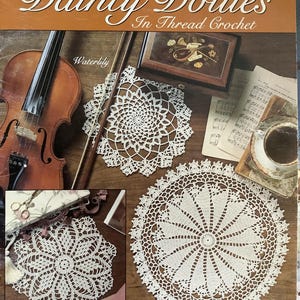 May include: A book cover titled "Dainty Doilies In Thread Crochet" with images of white crochet doilies. The cover includes a violin, a decorative box, sheet music, and the words "Waterlily", "Crown Jewels", and "Windmill".