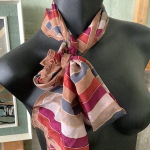 May include: A patterned silk scarf in a variety of colours, including red, purple, brown, and blue, draped over a black mannequin. The scarf features a geometric stripe design and is knotted at the neck. The edges of the scarf have a subtle border.