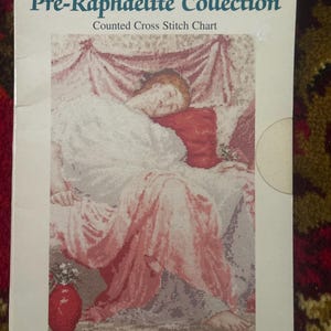 May include: A DMC Pre-Raphaelite Collection counted cross stitch chart booklet titled Jasmine. The cover features a printed illustration of a person resting on a bed with pink drapery, a red pillow, and a red vase with white flowers in the foreground.