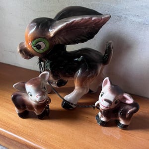 Puede incluir: Figura de burro de cerámica vintage con dos figuras de burro más pequeñas unidas por una cadena. El burro es principalmente negro y marrón con detalles blancos y ojos verdes grandes. Los burritos son marrones y blancos.