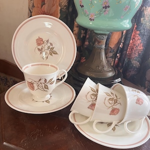 May include: A set of four white teacups and saucers with a floral design. The cups and saucers have a pink trim and feature a delicate floral pattern with brown leaves and pink roses.