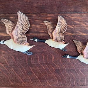 May include: Three decorative goose wall hangings in flight. Each goose has detailed wings with brown and tan feathers, a black head and neck, and a cream-colored body. The geese are arranged on a dark wood surface.