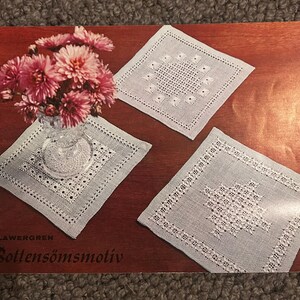 May include: Three square white lace doilies with intricate needlework patterns displayed on a reddish-brown surface. One doily supports a clear glass vase filled with pink chrysanthemum flowers. The text S. LAWERGREN BOTTENSÖMSMOTIV is visible.