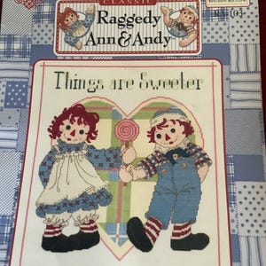 Può includere: Un modello a punto croce con Raggedy Ann e Andy che si tengono per mano e un lecca-lecca. Il testo "Things are Sweeter When Shared With a Friend" è visualizzato. Il design è ambientato su uno sfondo patchwork blu e bianco.
