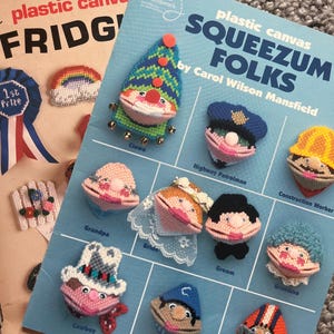 Op de afbeelding: Twee vintage plastic canvas knutselboeken worden getoond. Eén is getiteld "FRIDG" en de andere "SQUEEZUM FOLKS" door Carol Wilson Mansfield. Het boek "SQUEEZUM FOLKS" toont gestikte figuren, waaronder een clown, een politieagent en een cowboy.