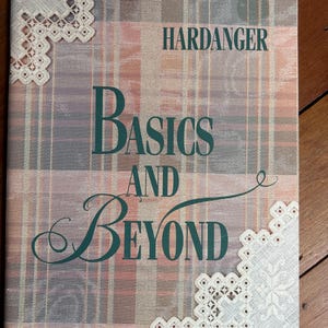 May include: A softcover book titled Hardanger Basics and Beyond by Janice Love. The cover features a muted plaid background in shades of pink, gray, and tan, decorated with white lace-like embroidery patterns in the top left and bottom right corners.