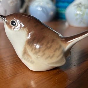 May include: A small ceramic bird figurine featuring a glossy finish with a cream-coloured chest and a brown back adorned with darker brown, dash-like markings. The bird has a pointed beak, a dark eye, and a short, upward-tilted tail.